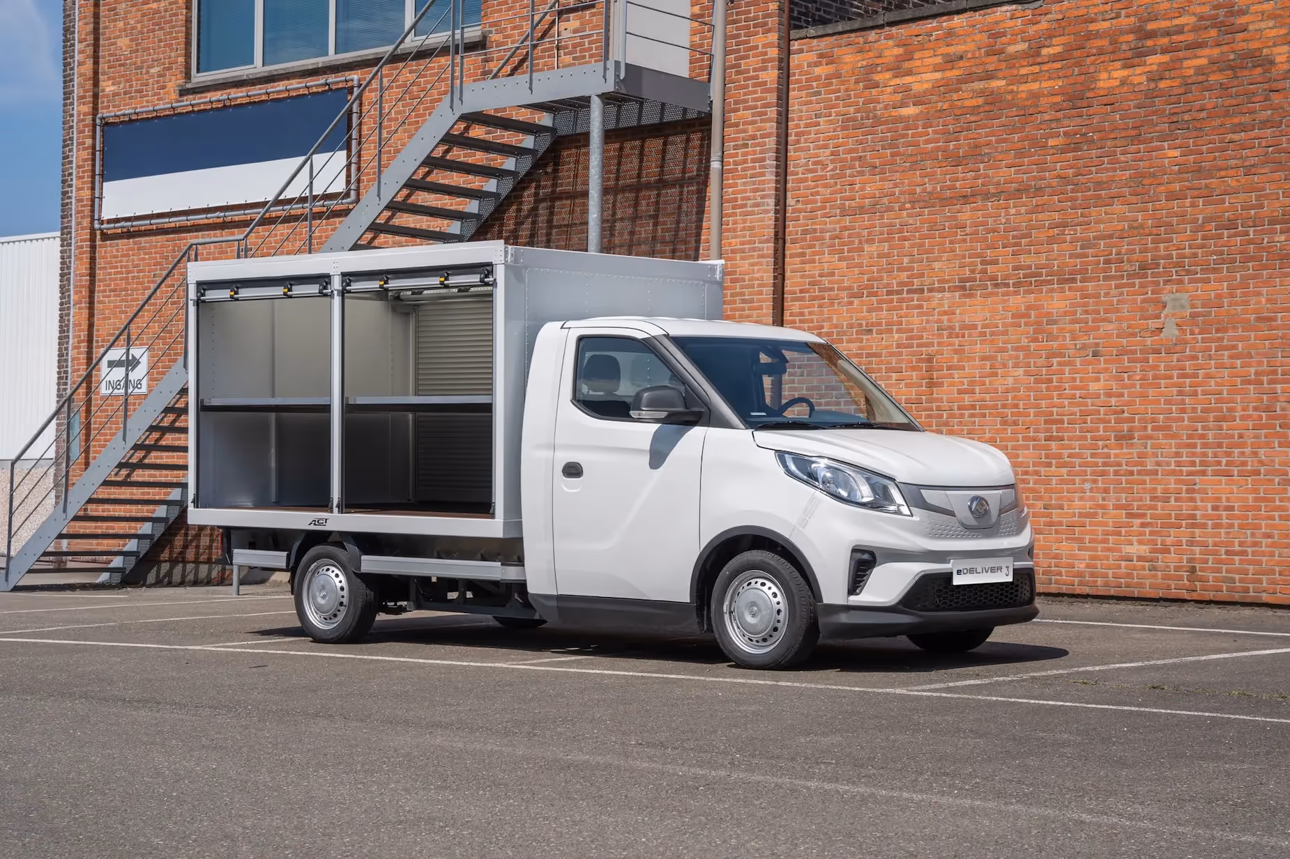 White electric delivery truck with open side compartments parked in front of a brick building with an exterior metal staircase.