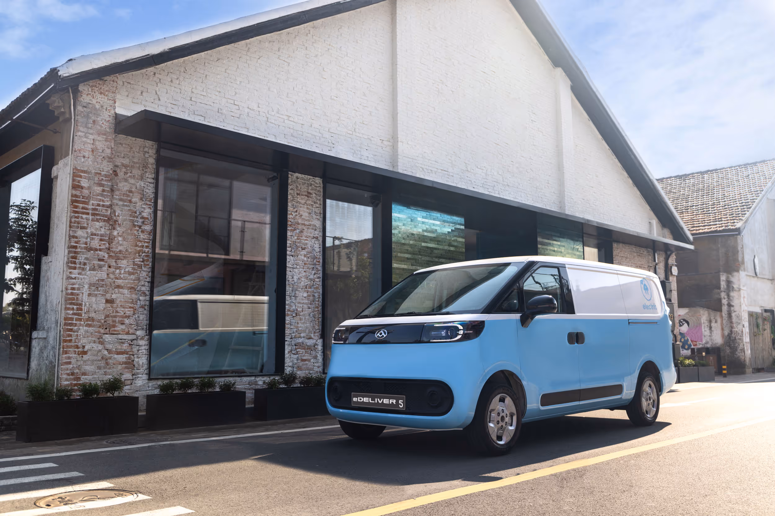 Light blue and white electric delivery van parked on a street beside a brick and white building.