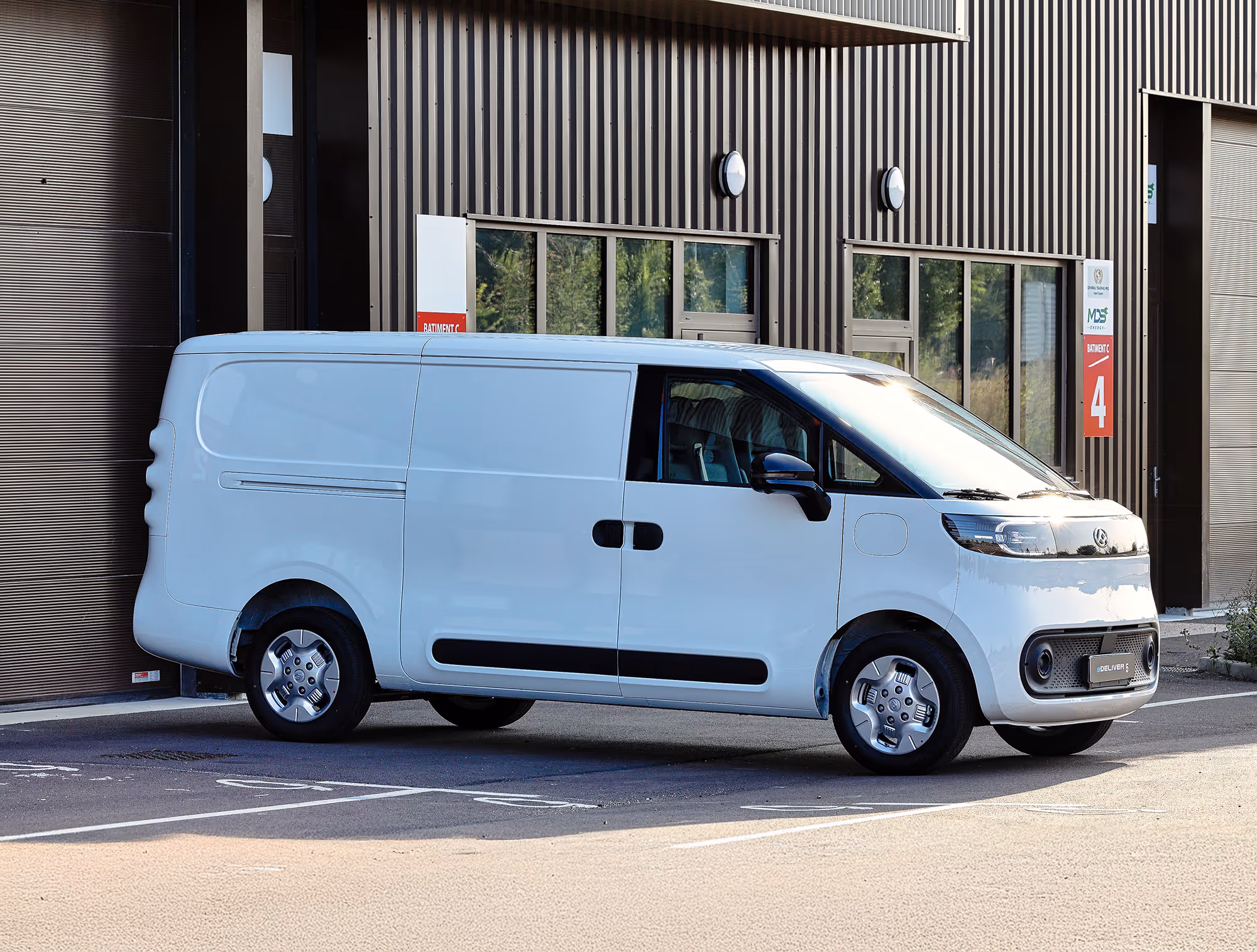 White compact electric delivery van parked in front of a commercial building with large windows and a red sign marked '4'.