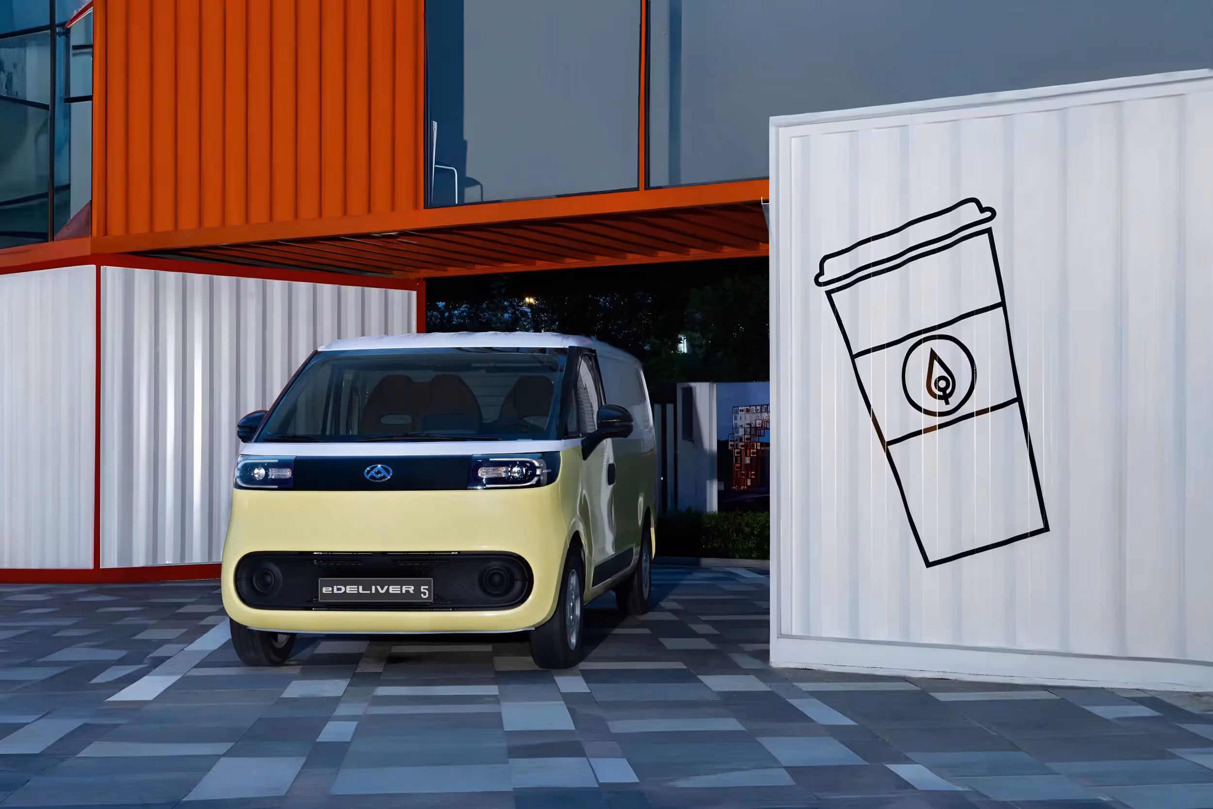 Front view of a compact yellow and white eDELIVER 5 electric van parked under a red shipping container structure at night.