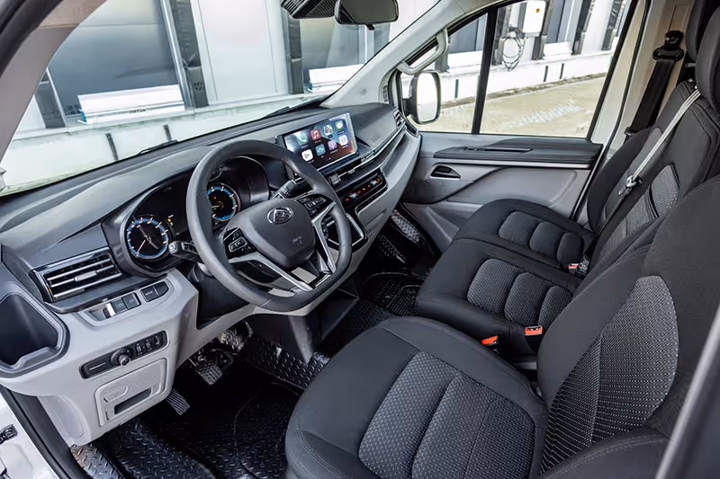 Interior view of a modern vehicle cabin showing black fabric seats, steering wheel with controls, digital dashboard, and touchscreen display.
