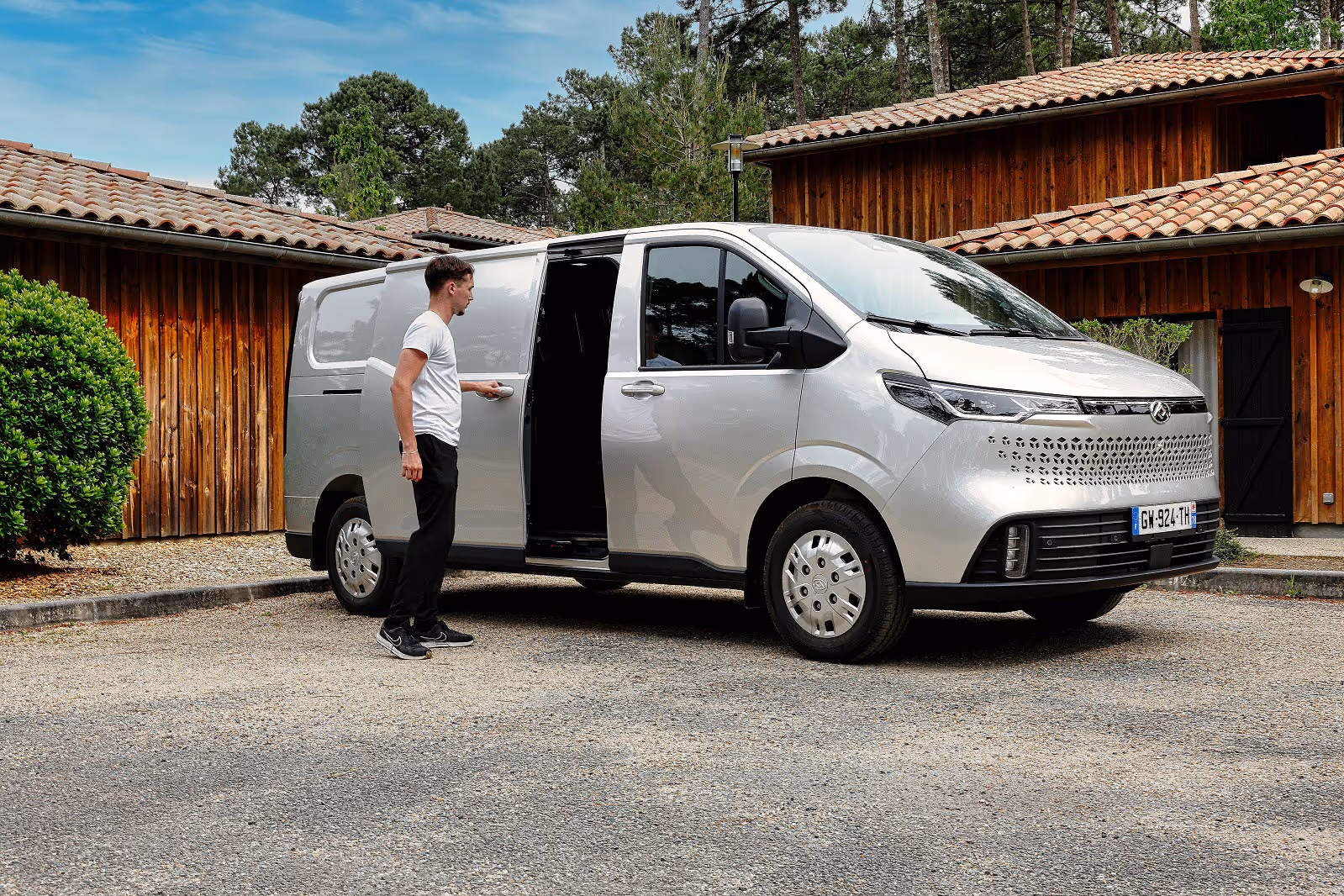 Man opening the sliding door of a silver van parked in front of a wooden building.