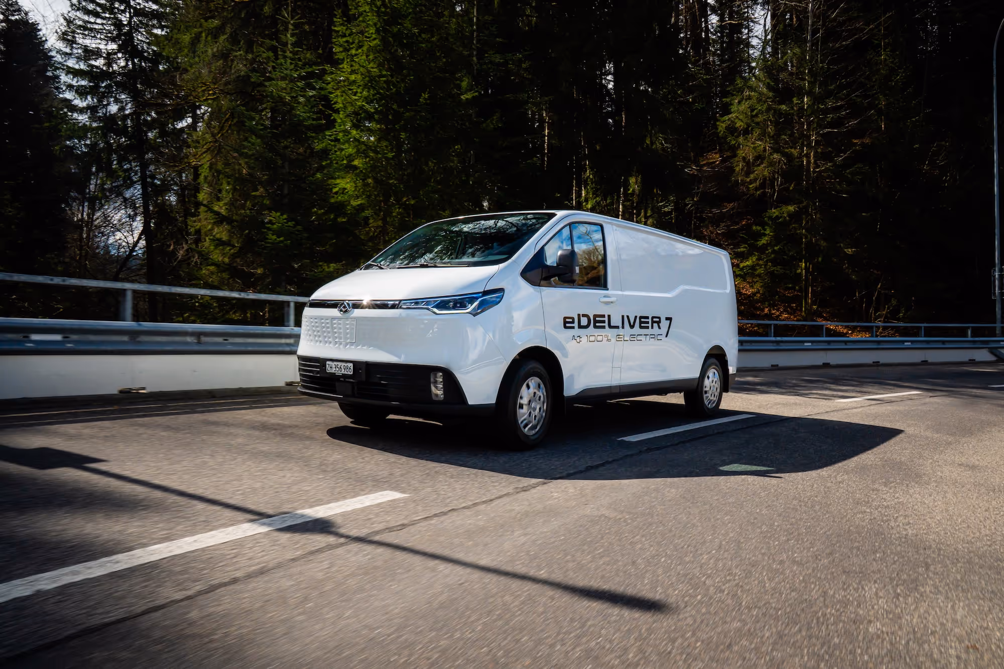 White electric delivery van labeled 'eDELIVER 7 100% ELECTRIC' driving on a forest-lined road.