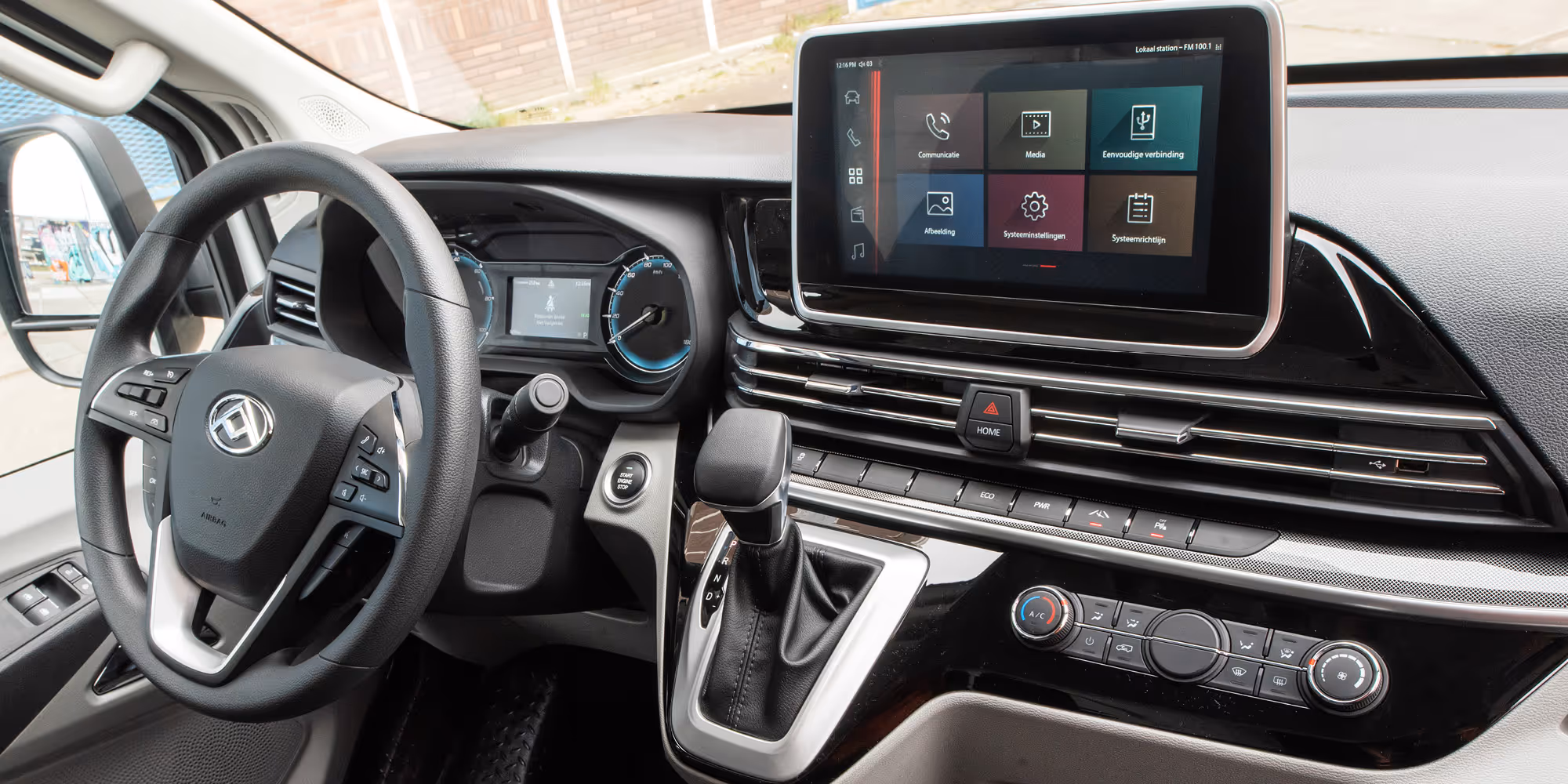Car interior showing steering wheel, gear shift, dashboard with digital display, and touchscreen infotainment system.