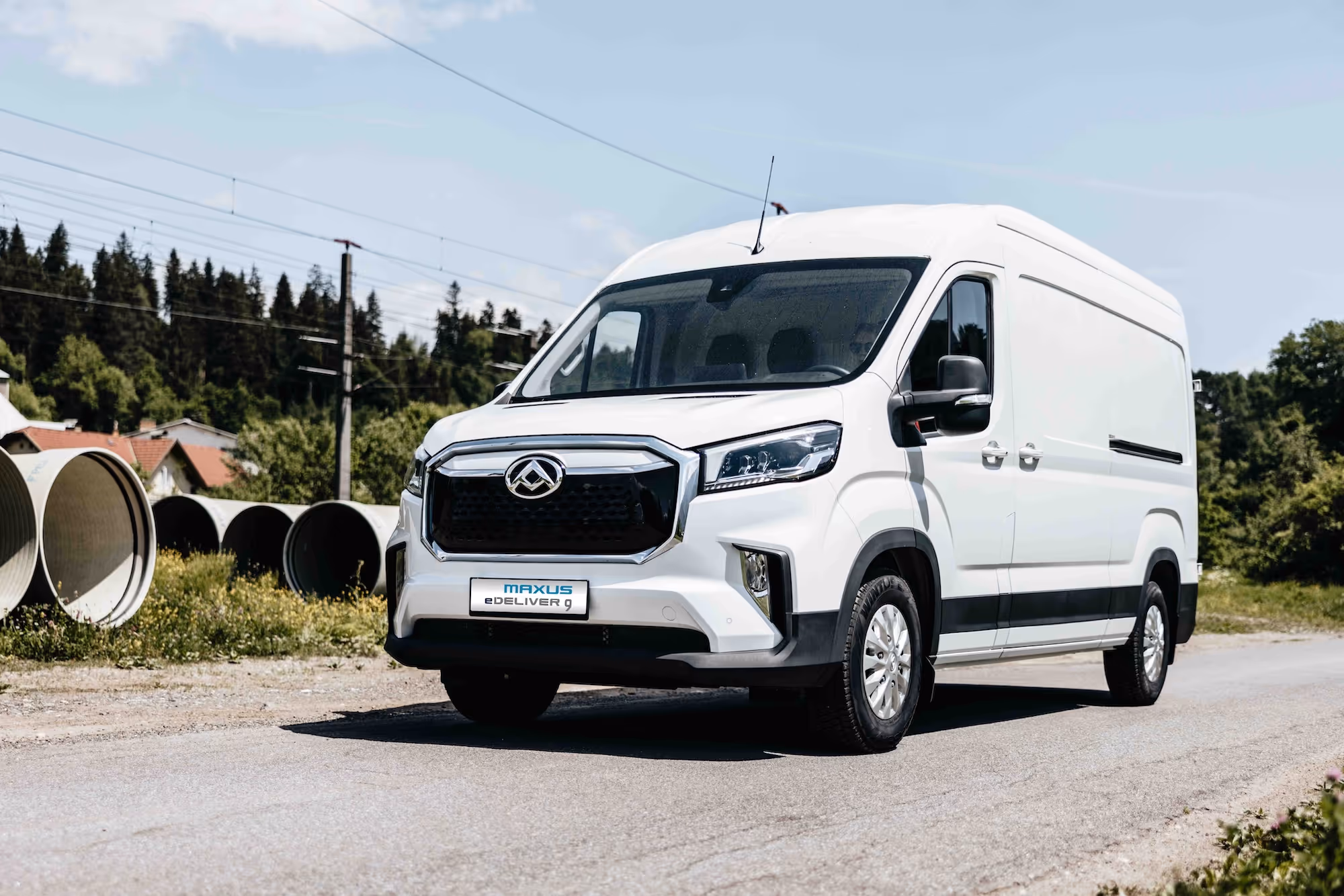White Maxus eDeliver 9 electric cargo van parked on a rural road with large concrete pipes and trees in the background.