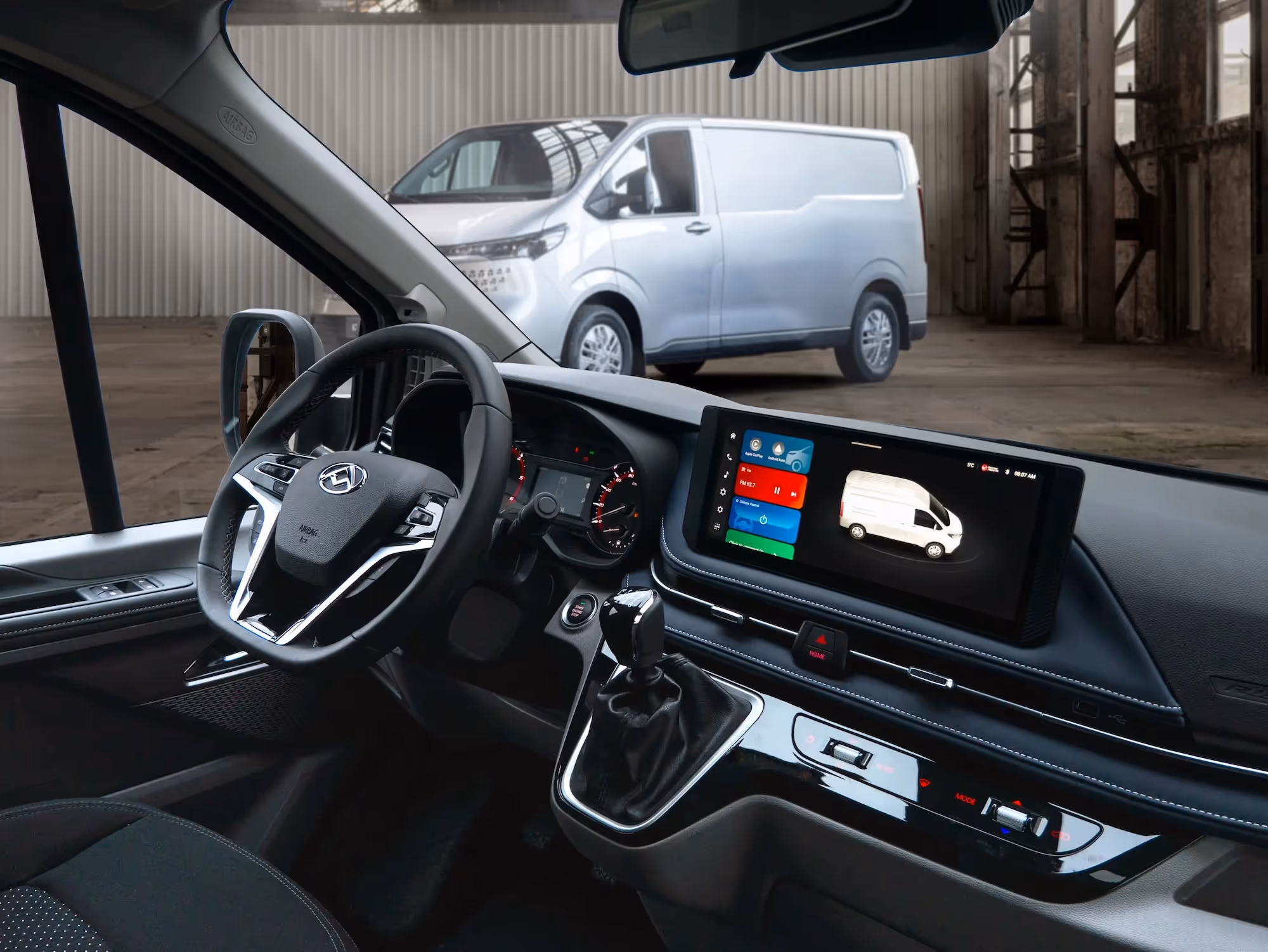 Interior view of a modern van's driver's seat showing the steering wheel, dashboard, gear shift, and infotainment screen with a van image displayed.