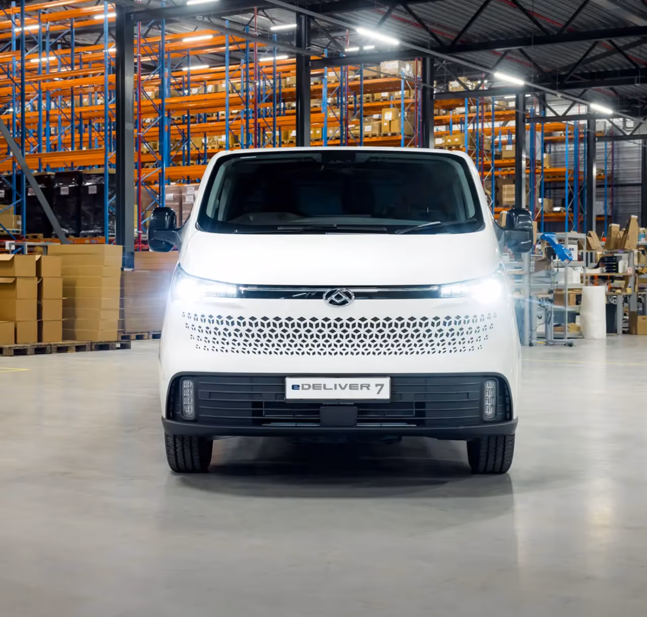 Front view of a white eDELIVER 7 electric delivery van with headlights on in a warehouse setting.
