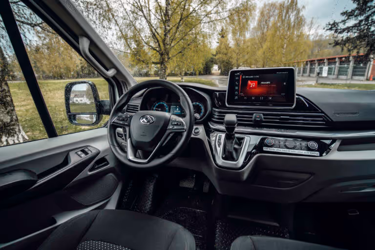 Interior view of a car's driver's seat, showing a steering wheel, dashboard with digital displays, a touchscreen infotainment system, and gear shift lever.