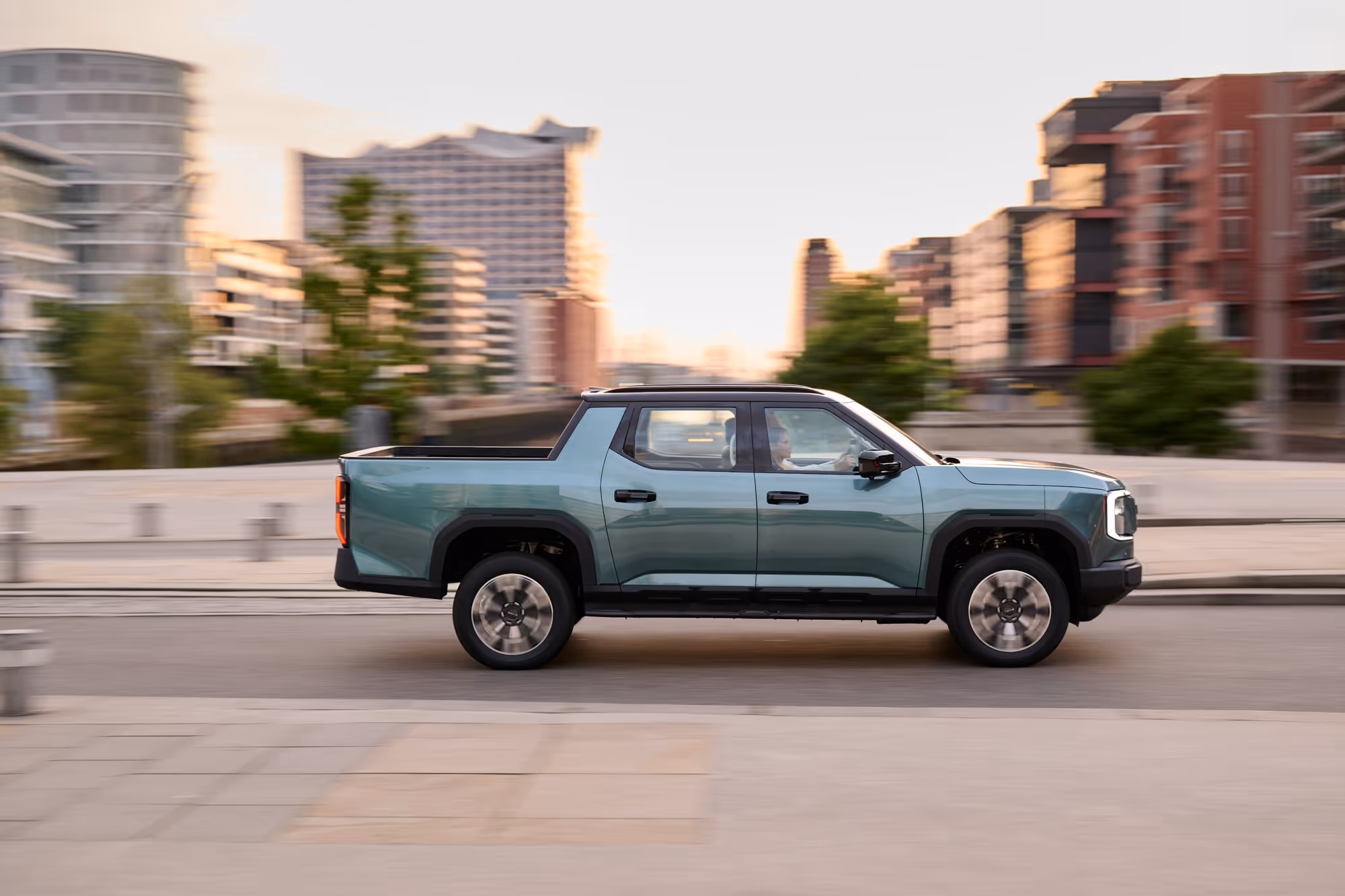 Side view of a teal electric pickup truck driving on an urban road with buildings in the background.