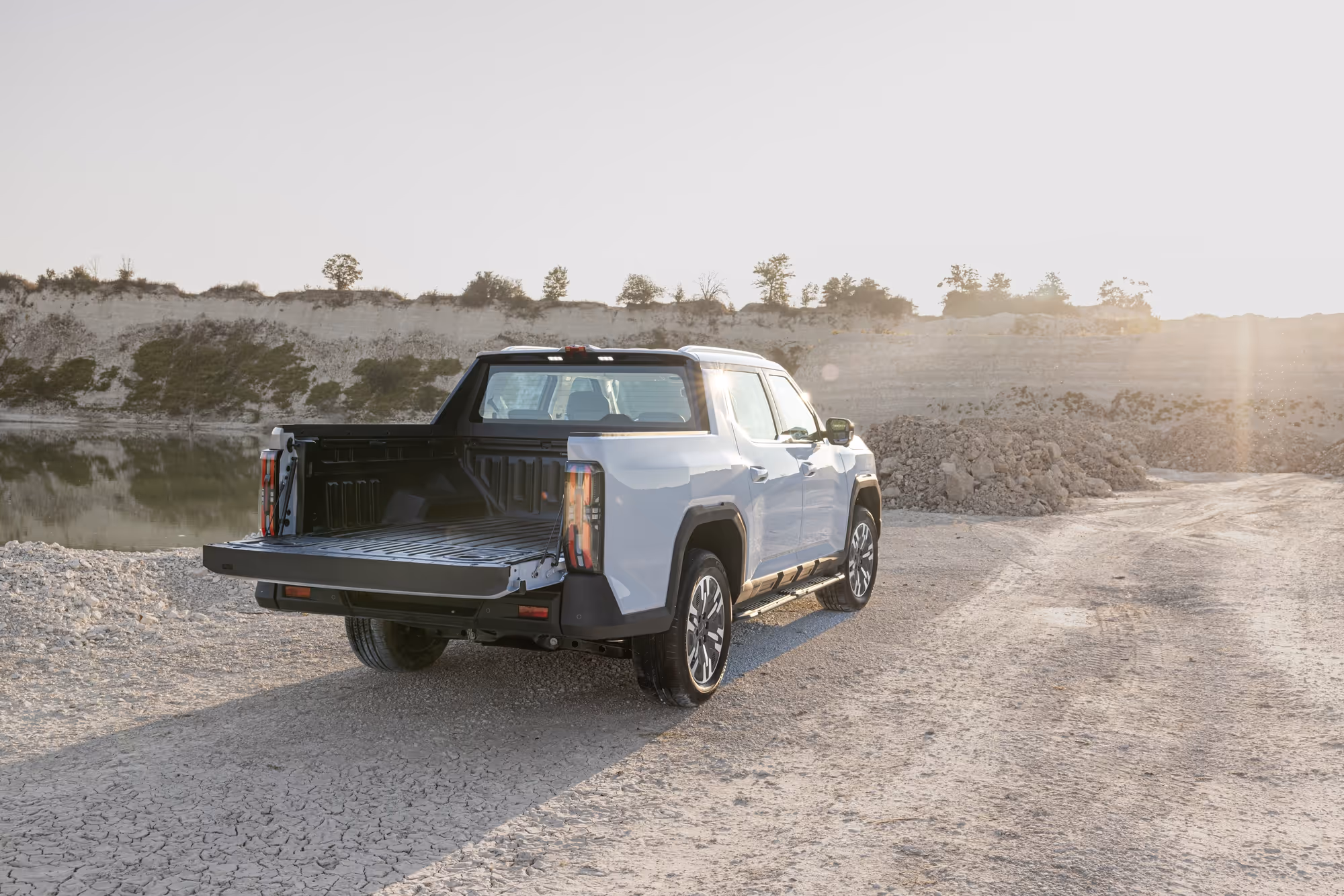 Modern white pickup truck parked on a dry, cracked dirt road near a rocky lake at sunset with its tailgate lowered.