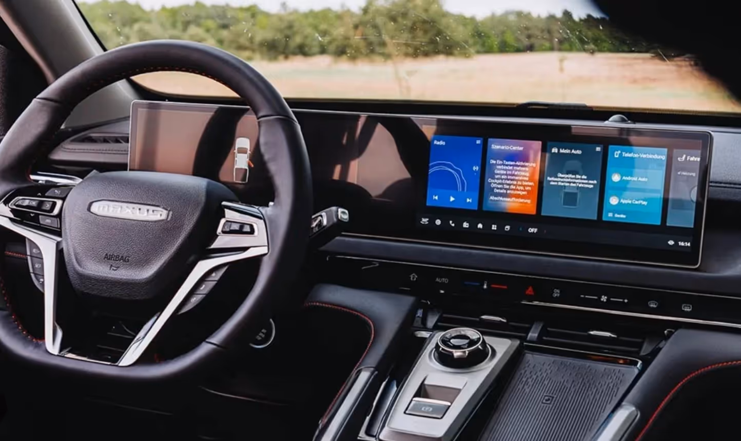 Modern car interior featuring a Maxus steering wheel and a wide digital dashboard display showing radio, vehicle settings, and phone connectivity options.