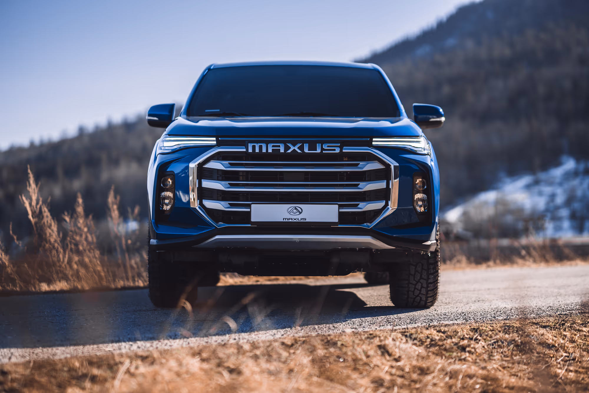Front view of a blue Maxus SUV parked on a mountain road with dry grass and blurred forest background.
