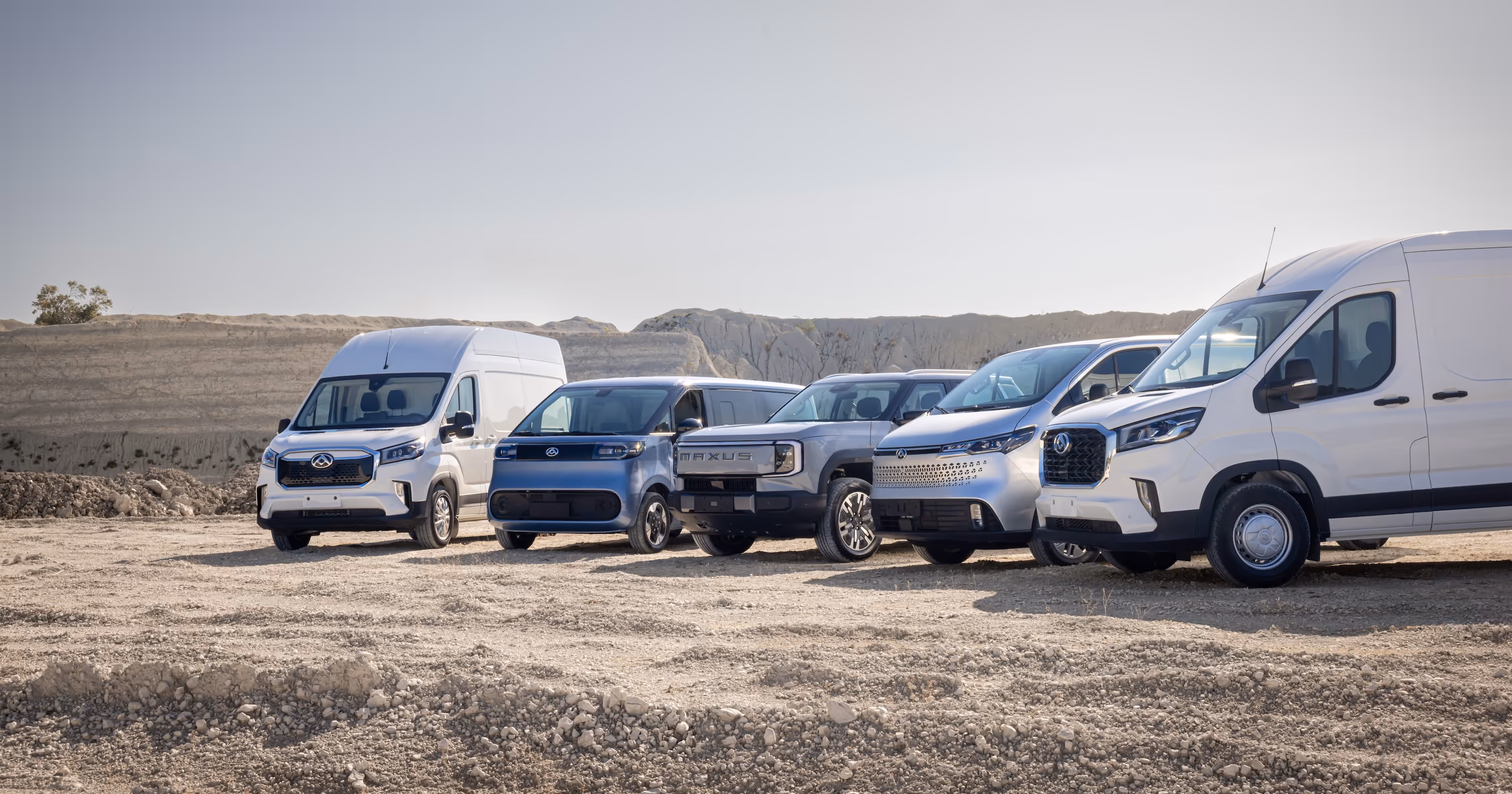 Five modern utility vehicles including vans and SUVs parked in a row on a dirt surface with hills in the background.