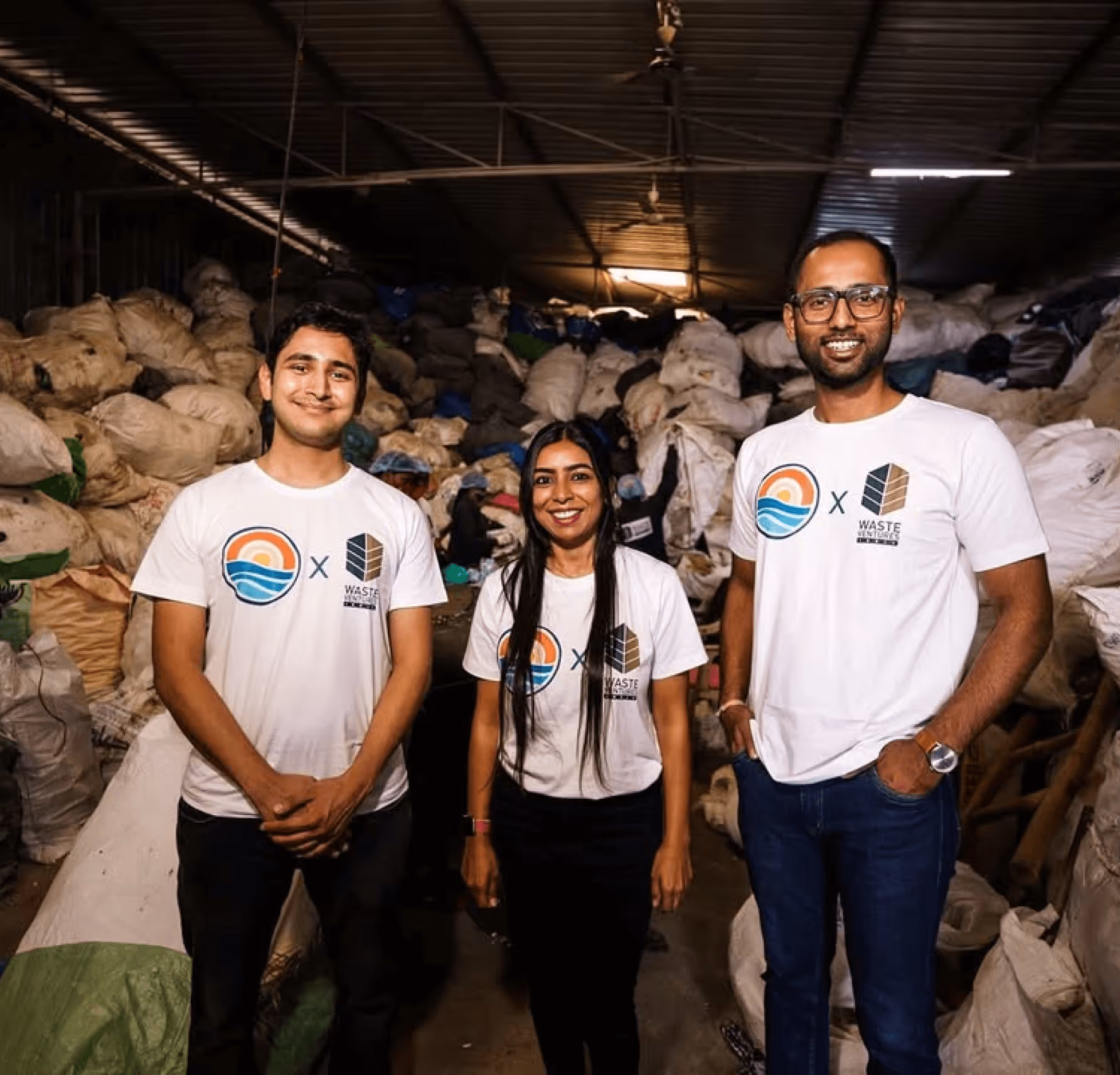 Three people wearing matching rePurpose Global × Waste Warriors t-shirts stand and smile in front of large piles of bagged plastic waste inside a recycling facility.