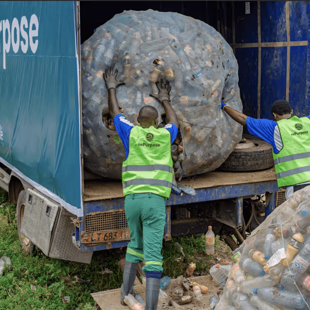 Two rePurpose Global workers in safety vests load a massive compressed bale of plastic waste onto a truck during a plastic recovery operation.