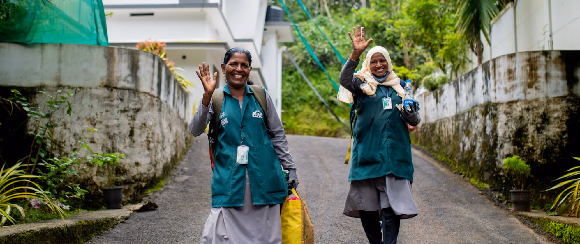 Two smiling waste workers in rePurpose vests wave at the camera while walking up