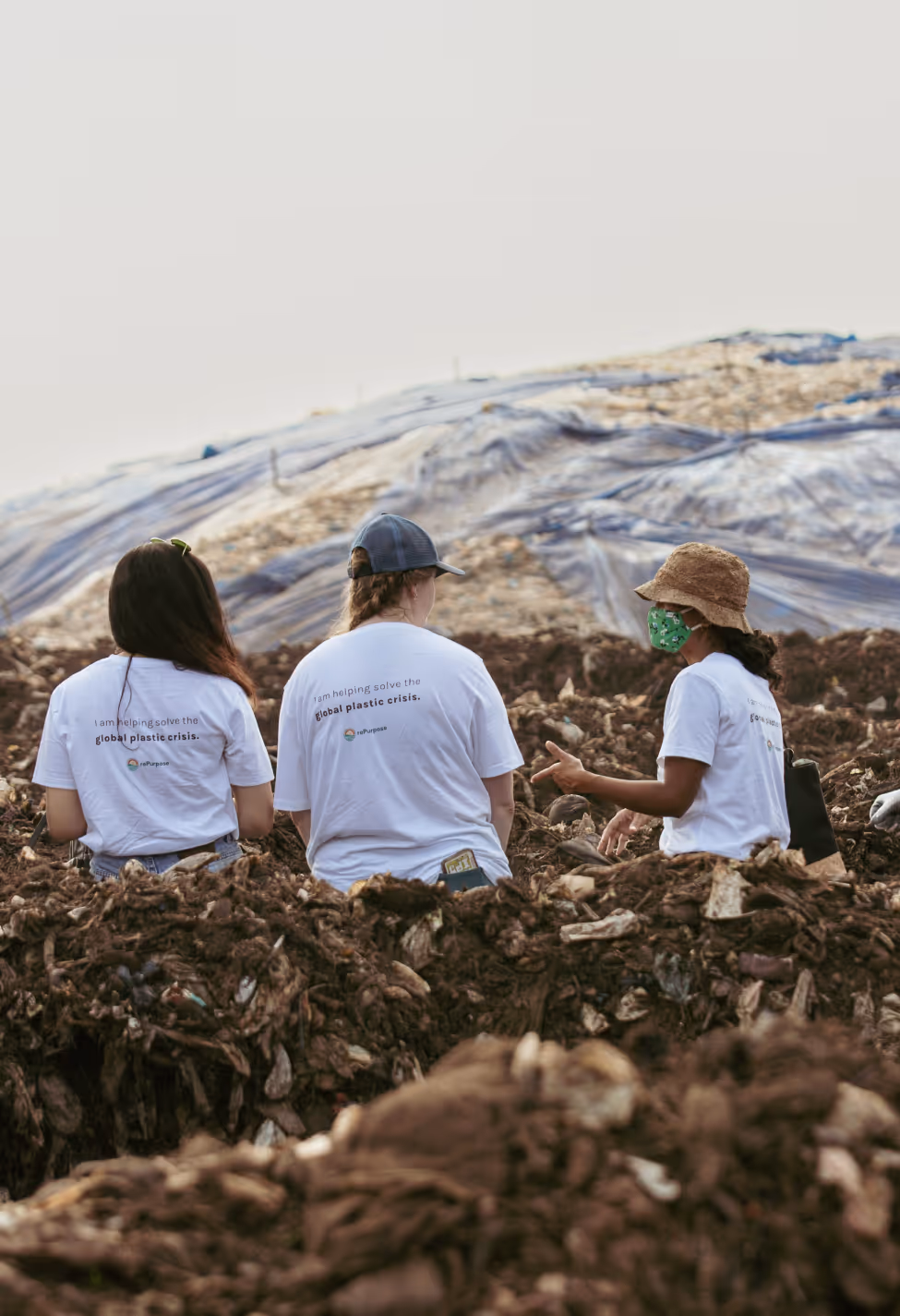 Three rePurpose Global volunteers wearing branded white T-shirts sit at a large landfill site, discussing plastic waste solutions as they face a plastic-covered hillside.