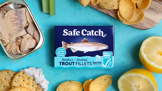 Box of Safe Catch skinless smoked trout fillets surrounded by crackers, lemon halves, celery sticks, and an open can of trout fillets on a turquoise surface.