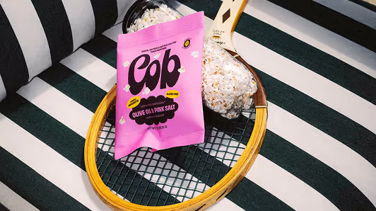 Pink bag of Cop Olive Oil Pink Salt popcorn resting on a wooden tennis racket, with popcorn visible in a black bowl, all placed on a green and white striped surface.