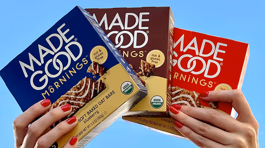 Hands with red nail polish holding three colorful boxes of Made Good Mornings soft baked oat bars against a blue sky.