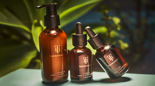 Three amber glass skincare bottles with black caps on a surface, labeled True Botanics, against a blurred green plant background.