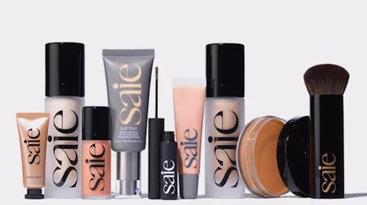 Various Saie brand makeup products including foundation, concealer, tinted moisturizer, mascara, and a makeup brush arranged side by side.