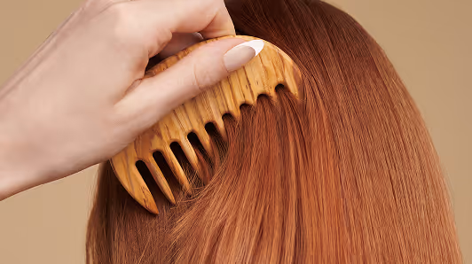 Hand with manicured nails combing smooth, straight reddish-brown hair with a wooden wide-tooth comb.