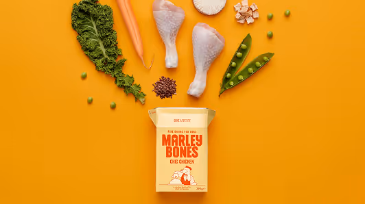 Marley Bones chic chicken dog food box with scattered ingredients including kale, carrot, chicken drumsticks, mushrooms, peas, and flax seeds on an orange background.