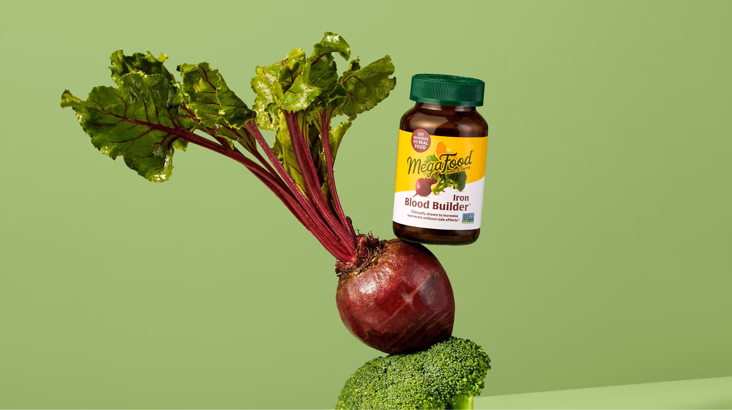 A bottle of MegaFood Iron Blood Builder supplement balanced on a beetroot, which is placed on a broccoli floret, all against a green background.