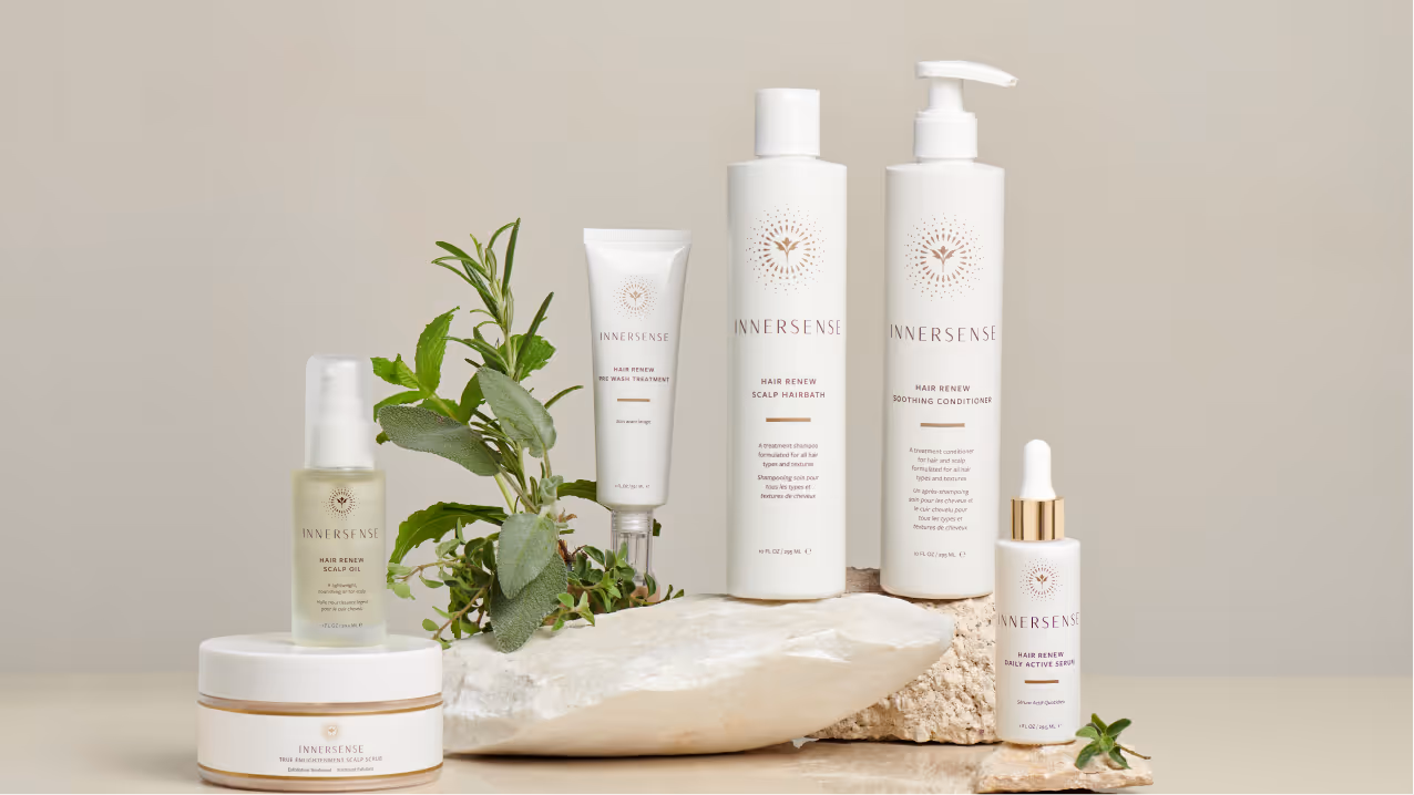 A set of six Innersense hair care products arranged with green herbs and white stones on a beige background.