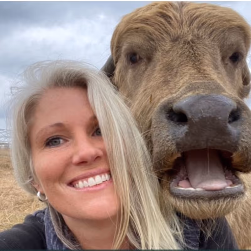 Smiling woman with blonde hair taking a close-up selfie with a brown cow outdoors.