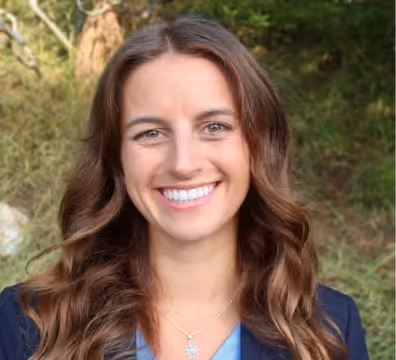 Smiling woman with long wavy brown hair wearing a blue blazer and necklace, outdoors with greenery in the background.