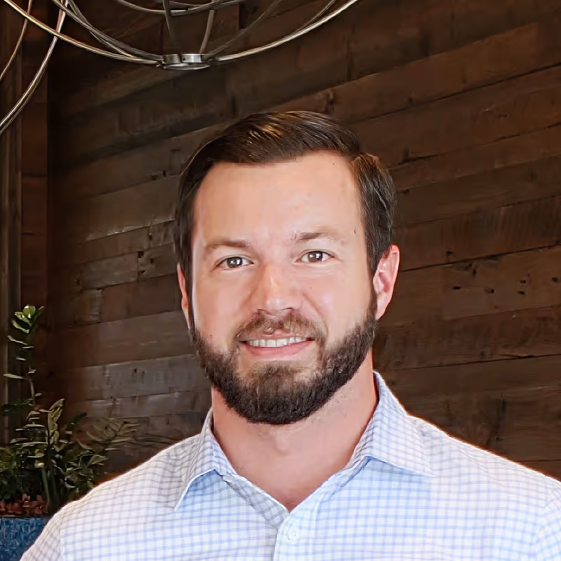 Smiling man with beard in a checkered shirt standing in front of wooden paneling and plants.