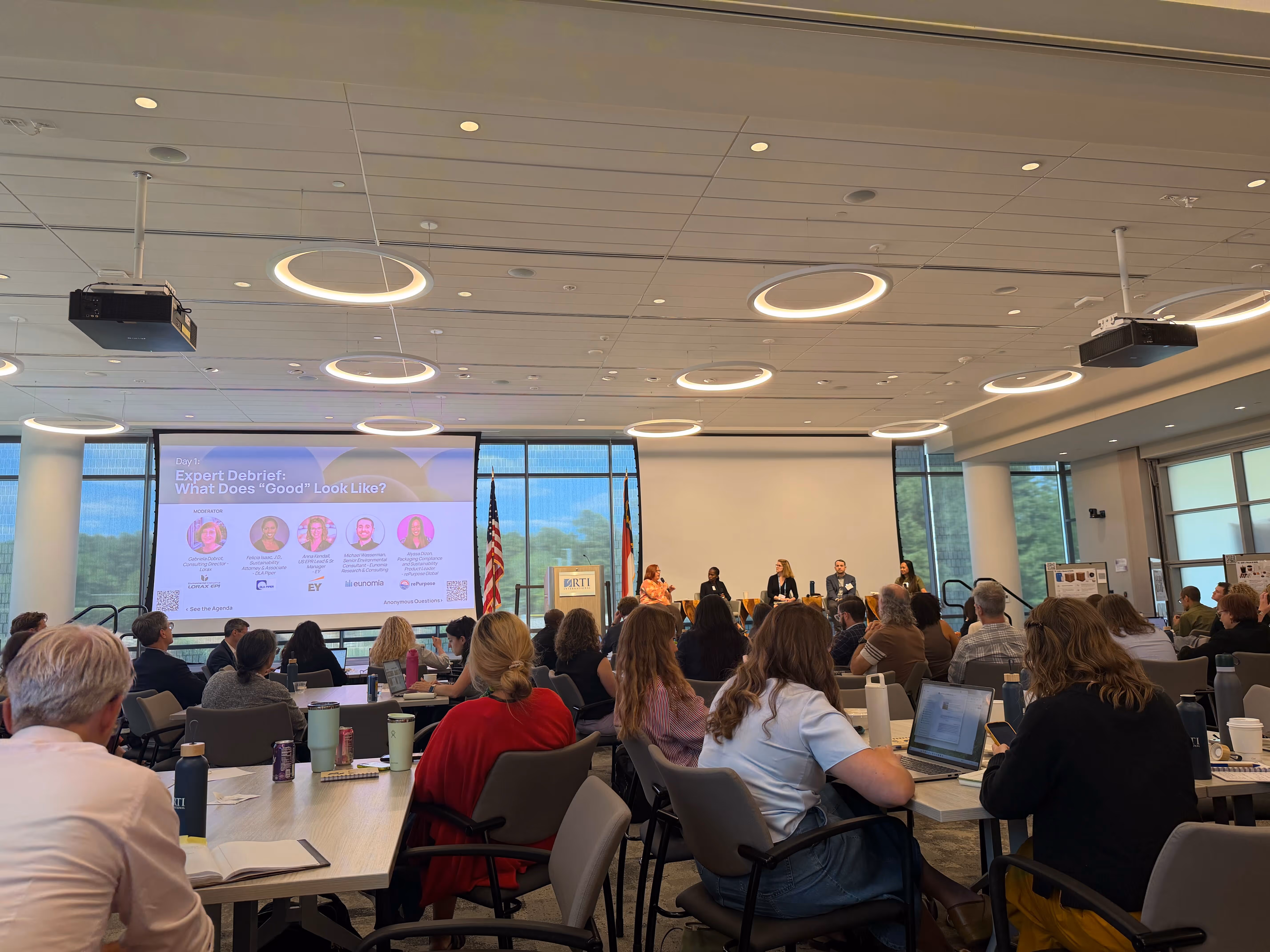 Audience at a conference room panel discussion with presentations on screens and round ceiling lights.
