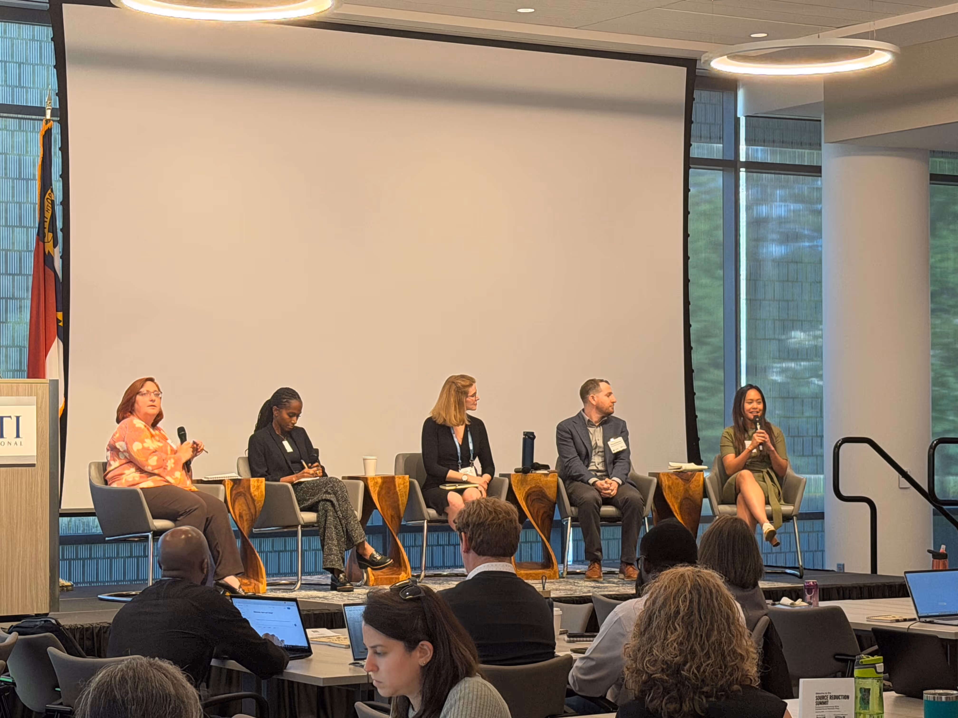 Five panelists seated on stage in front of a large screen speaking to an audience.