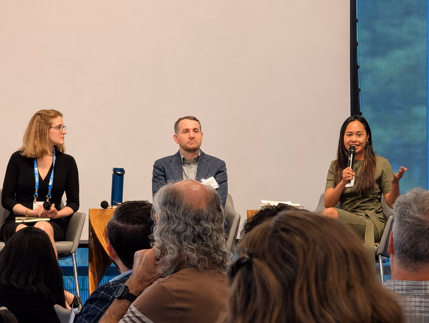 Three panelists seated on stage, one woman speaking into a microphone, audience visible from behind.