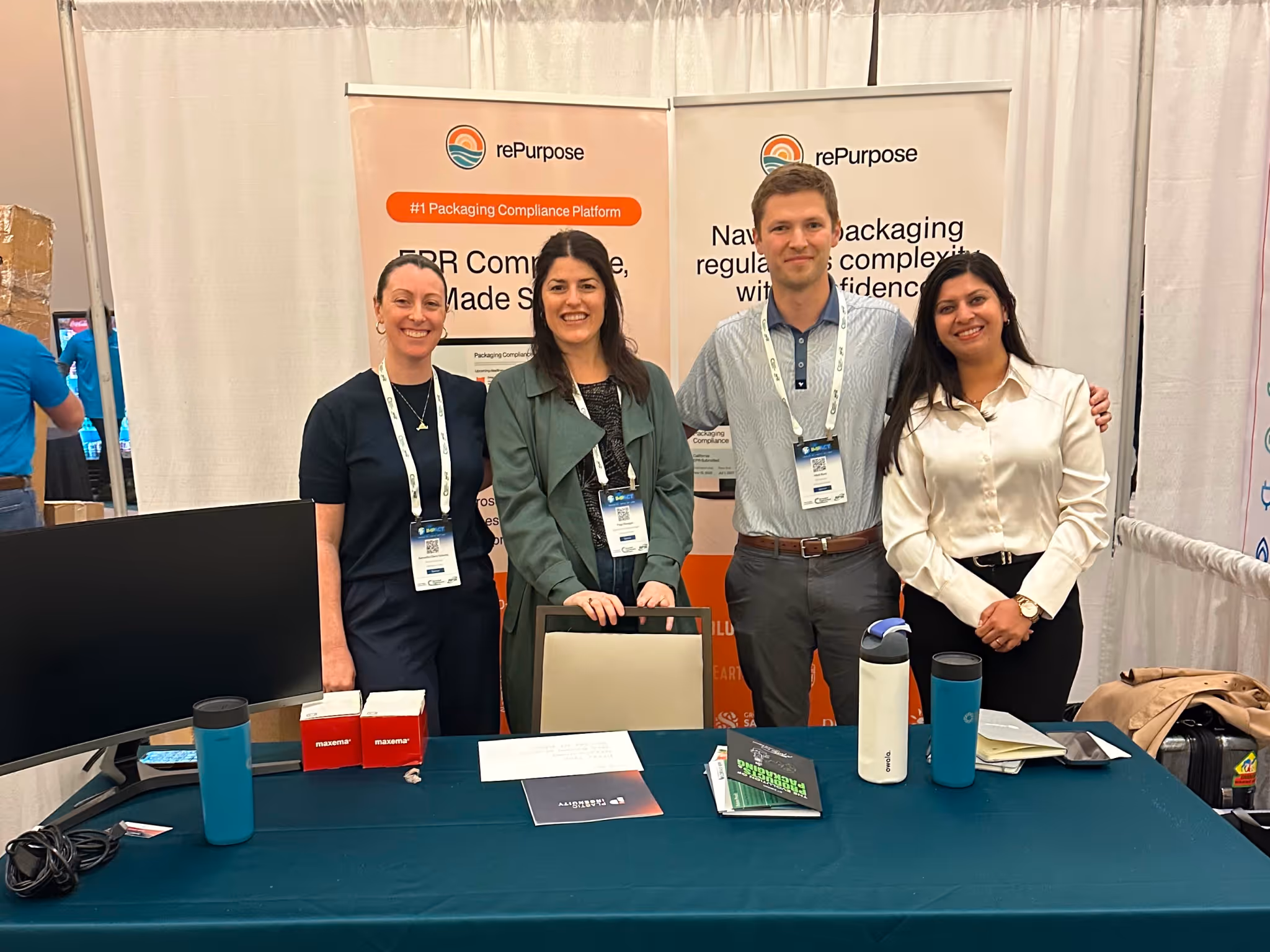 Four smiling people standing behind a table at a rePurpose trade show booth with banners.
