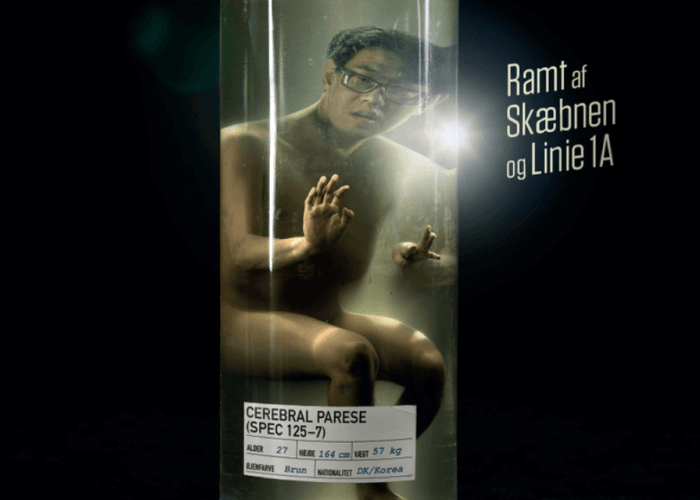 A young man with glasses, appearing naked and confined inside a large glass jar or cylindrical container. He has a concerned or anxious expression and is pressing his hands against the glass as if trying to get out. There is a bright light reflection on the glass. The background is dark, and to the right, Danish text reads "Ramt af Skæbnen og Linie 1A." A label on the jar reads:, CEREBRAL PARESE (SPEC 125-7), ALDER: 27  HØJDE: 164 cm  VÆGT: 57 kg, ØJENFARVE: Brun  NATIONALITET: DK/Korea.