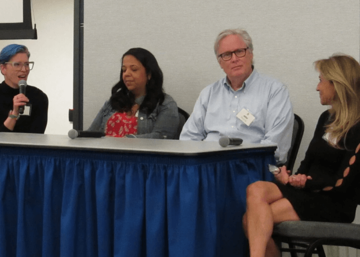 Three women and one man sitting together at a table, during a panel. The woman on the far left is speaking into a microphone.