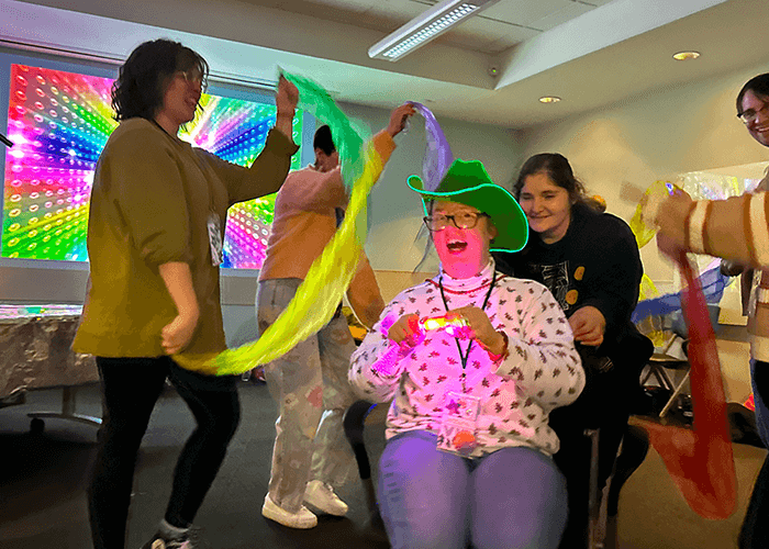 A person in a wheelchair, wearing a long sleeve shirt, jeans, and a green cowboy hat. They are surrounded by four people holding scarves, presumably dancing with them around said person.