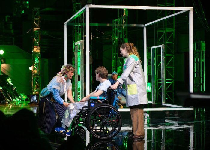 Two women and one young boy in a wheelchair on a green live performance set.