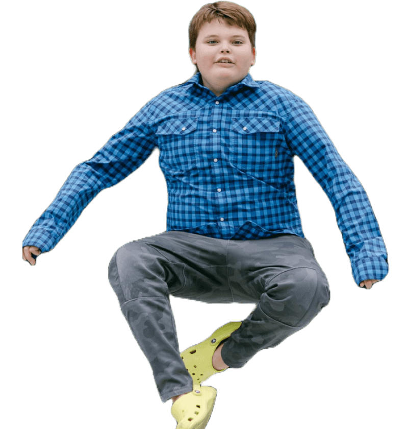 A young boy with short brown hair, wearing a darker blue plaid button up, camo pants, and light green crocs. He is looking at the camera, mid-air. The background is orange.