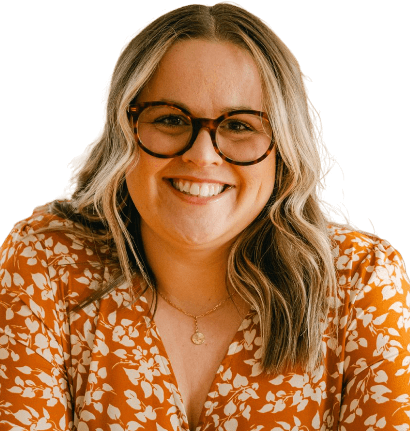 A white woman with medium length curled dirty blonde hair, wearing glasses and an orange floral top. She is looking at the camera, smiling. The background is a dusty rose.