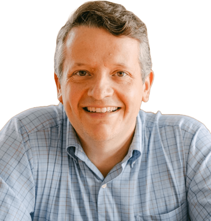 A white man with short brown hair, wearing a light blue plaid button up. He is looking at the camera, smiling. The background is blue.