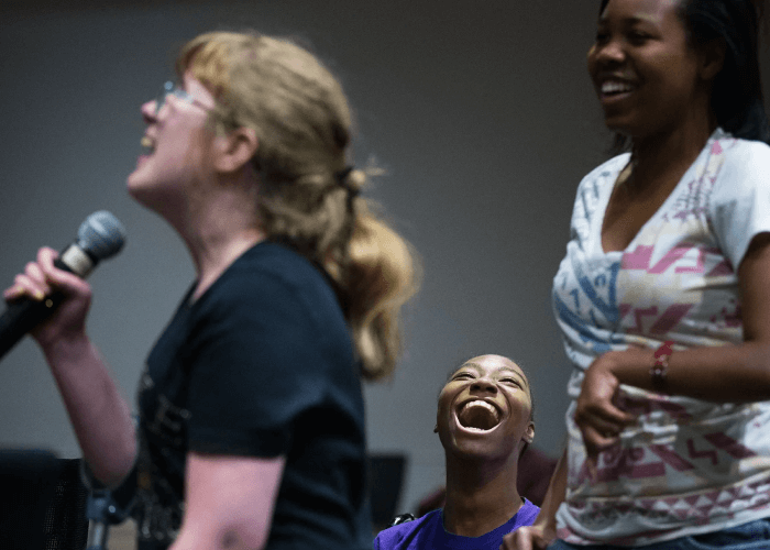 A white woman with her hair pulled back, wearing glasses, is speaking into a microphone. One Black woman is sitting behind her, head back in laughter. Another Black woman stands beside her, smiling.