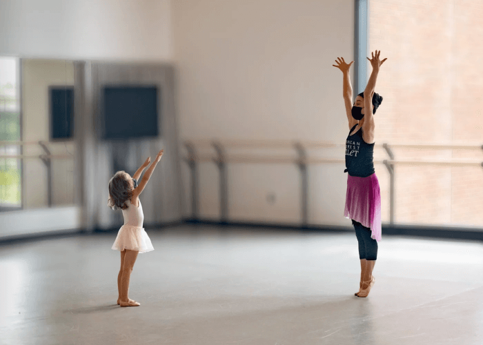 A small child and an adult are ballerinas dancing together, arms in the air while standing on their tip toes and feet.