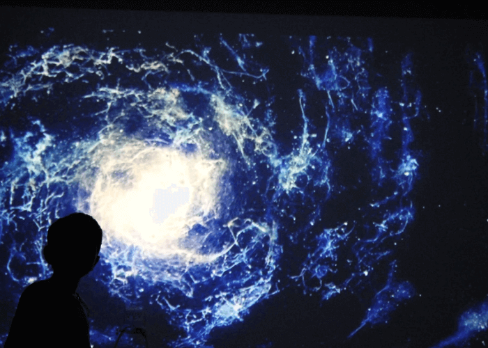 Silhouette of a person in front of a swirling blue and white cosmic pattern that looks like a galaxy. The bright center radiates outward with intricate lines and shapes, resembling clouds or nebulae in space. The background is dark, making the illuminated patterns stand out vividly. The scene has a sense of awe and wonder, capturing the vastness and beauty of the cosmos.