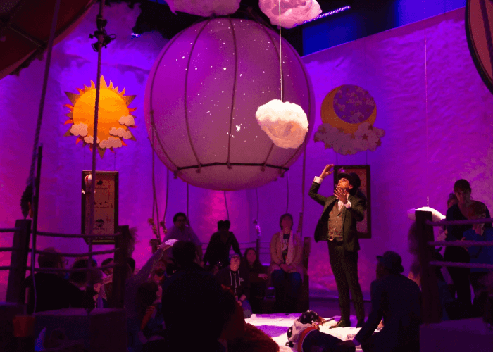 A whimsical indoor scene with purple lighting, featuring a ringmaster-like performer standing in the center of a ring, surrounded by an audience. Above, there are large hanging decorations: a glowing sun, a starry moon, fluffy clouds, and a giant balloon-like globe with twinkling star lights. The setting feels magical and theatrical, evoking a sense of wonder and imagination, with children and adults watching the central figure's performance.