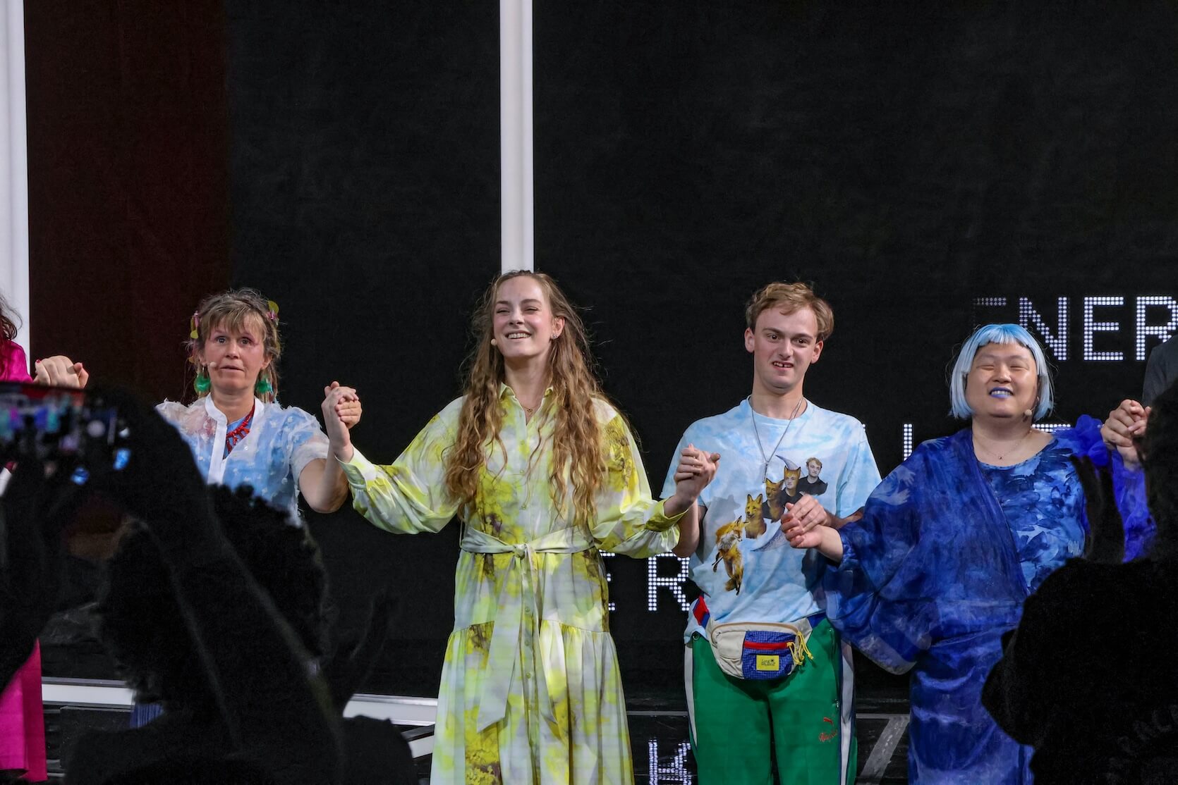 Four people standing on a stage, holding hands and forming a line. The group is taking a bow. The person second from the left, a young woman with long wavy hair, is smiling, wearing a light yellow and white tie-dye dress. The person next to her on the right, a young man with a fanny pack, is wearing a T-shirt with animal and person graphics and green pants. On the far right is a person with short light blue hair, matching blue lipstick, and a blue patterned dress, and is smiling with closed eyes, as they are Blind. The person on the left has colorful hair accessories and earrings, wearing a see-through blue garment. Some audience members in the foreground are raising their hands and capturing the moment on a phone. Behind them, part of a screen with white text on a black background is visible.