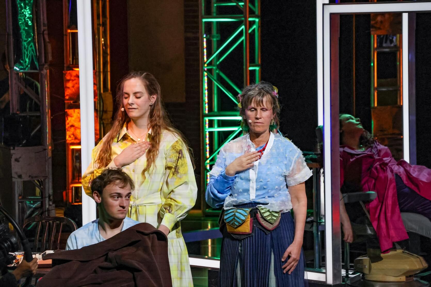 Theater scene with three people in the foreground and one person in the background. The two women in the center are standing next to each other with their eyes closed and their right hands placed on their chests, singing. The younger woman has long light brown hair and is wearing a light yellow checkered dress. The older woman has short hair, a blue shirt with transparent sleeves, and a multicolored belt bag. In front of them, a young man is sitting, covered with a brown blanket, looking off to his side with a neutral expression. In the background, there is another person reclining in a red outfit, almost lying down with their head back and arm extended.